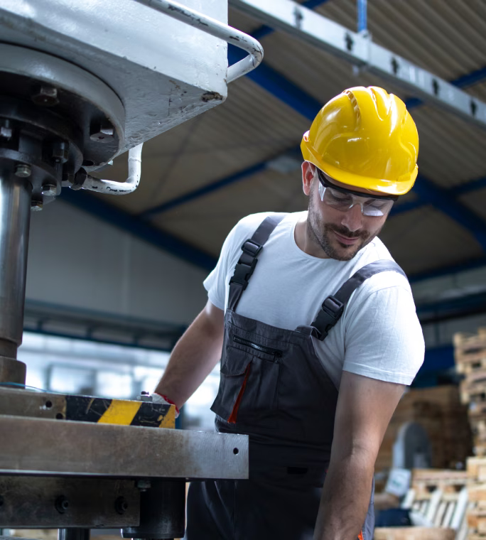 Opérateur portant un casque jaune et des lunettes de protection, penché près d’une machine industrielle.