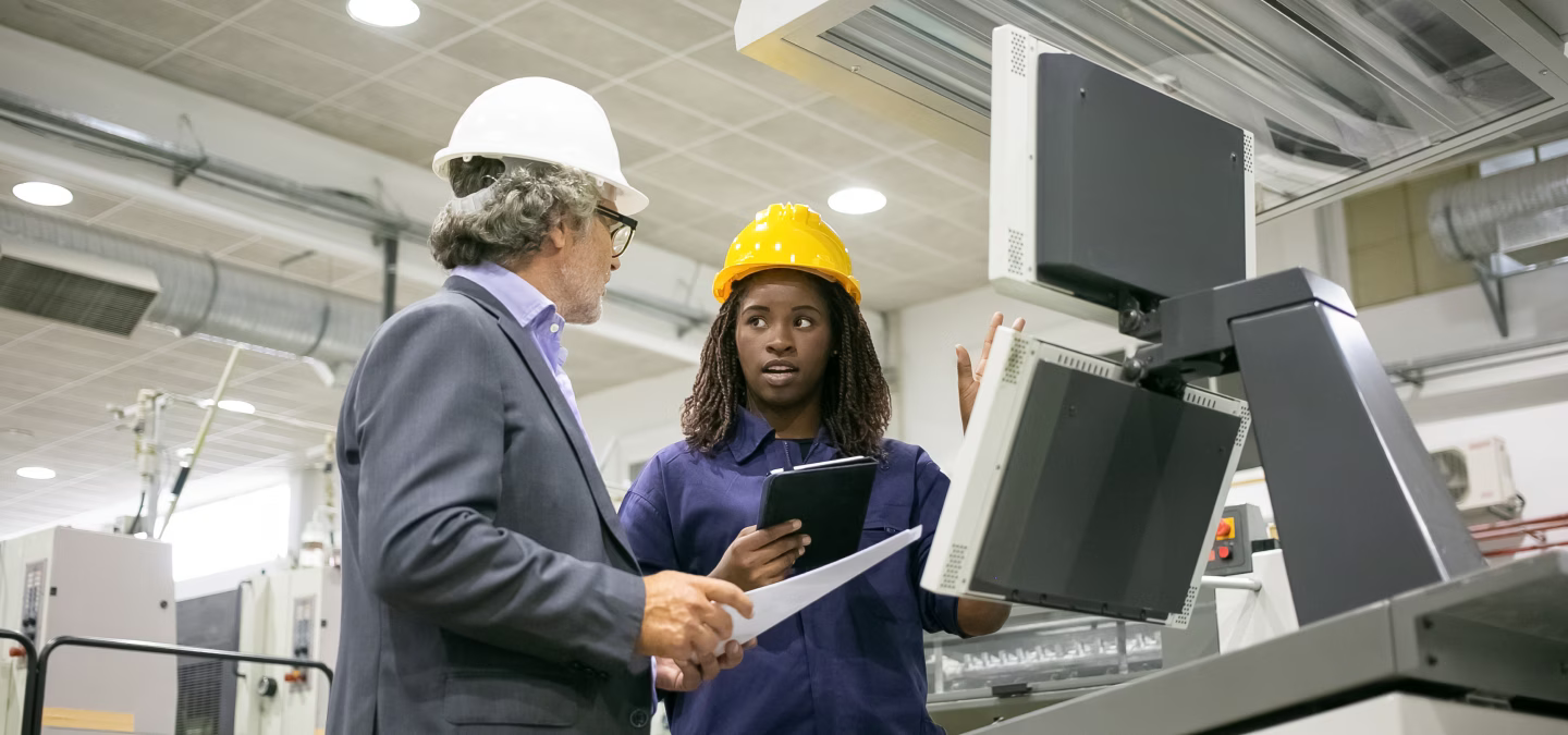 Deux personnes portant des casques échangent près d’écrans industriels, avec une tablette et un document papier.